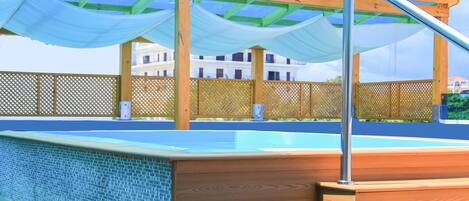 Une terrasse en bois avec une piscine, une pergola recouverte d’un tissu bleu et une clôture en bois.