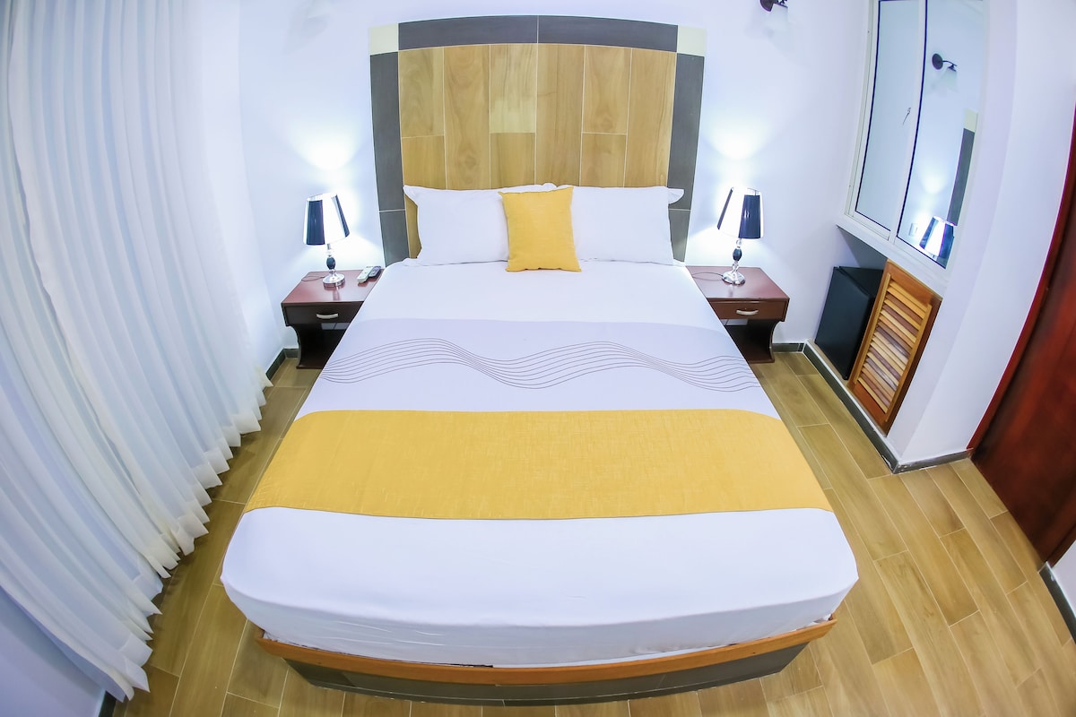 Ein ordentlich gemachtes Bett mit Holz-Kopfteil, zwei Nachttische mit Lampen, einem Spiegel und einem kleinen Schrank.