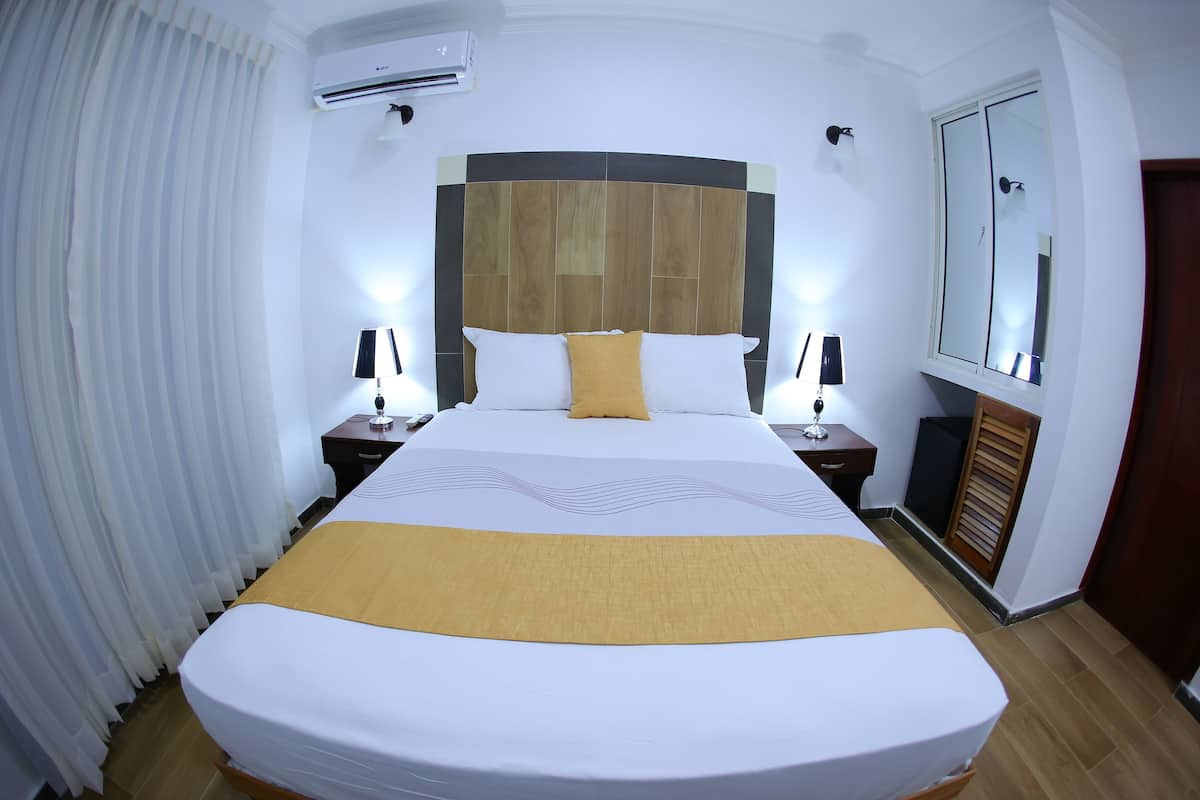 Een hotelkamer met een groot bed, een houten hoofdbord, twee nachtkastjes met lampen, een spiegel en een raam met jaloezieën.