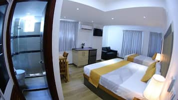 A hotel room with a bed, bedside table, lamp, and a desk with a chair. There is a shower area with a glass door and a toilet visible in the corner.