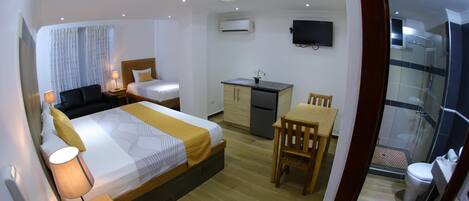 Habitación de hotel pequeña con cama, mesita de noche, lámpara, escritorio, silla y una pequeña cocina.