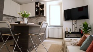 Una cocina moderna con una isla central, un comedor con sillas, un sofá con cojines, un televisor de pantalla plana y una ventana con luz natural.