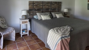 Un dormitorio con una cama, dos mesitas de noche, una lámpara, una silla y un pequeño estante con libros.