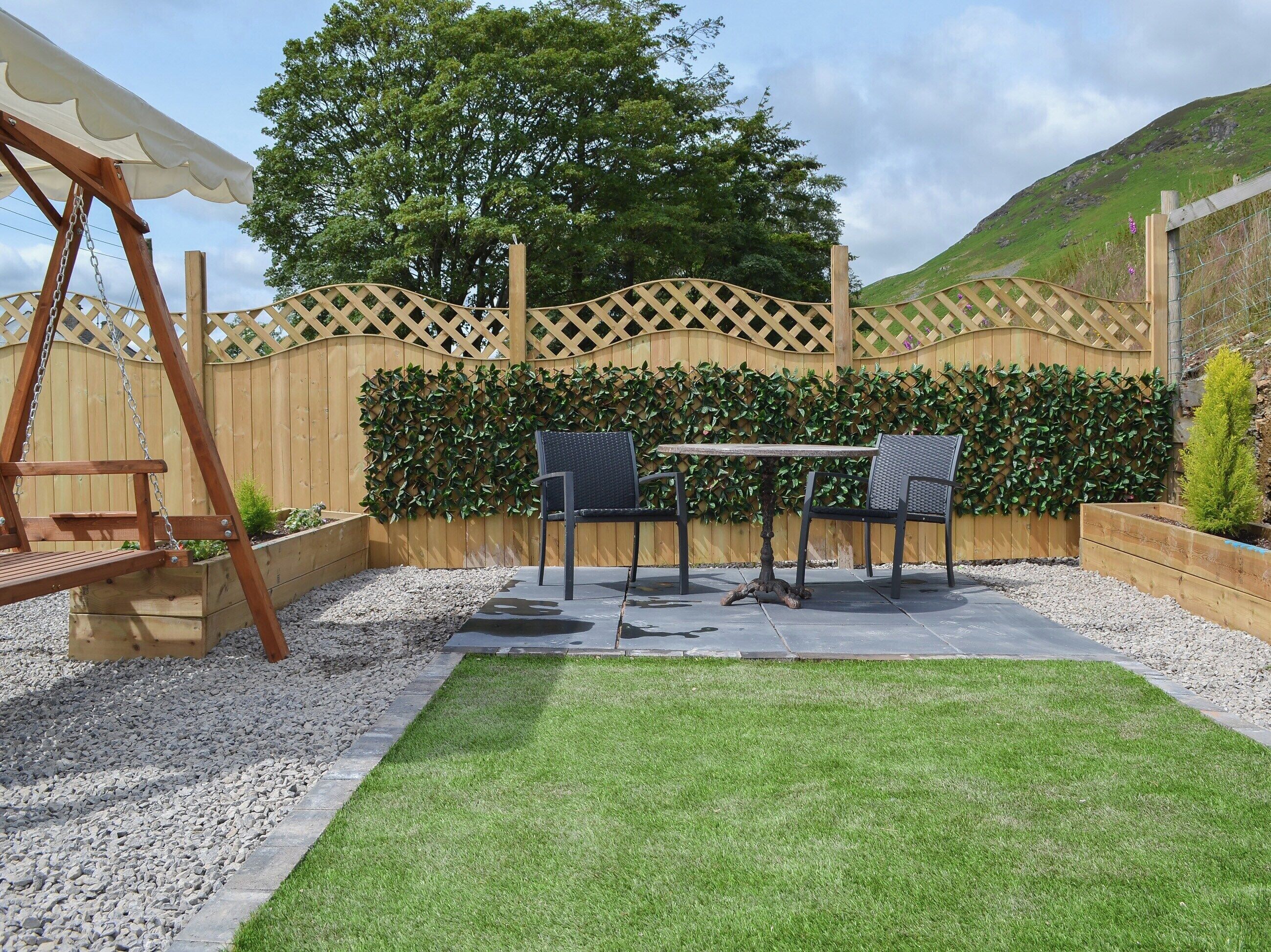 A patio with a swing set, two chairs, a table, and a decorative hedge wall.