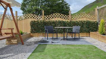 A patio with a swing set, two chairs, a table, and a decorative hedge wall.