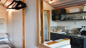 A modern bedroom with a large mirror, a bed with striped bedding, and a view into a kitchen with a microwave, sink, and various kitchen items.