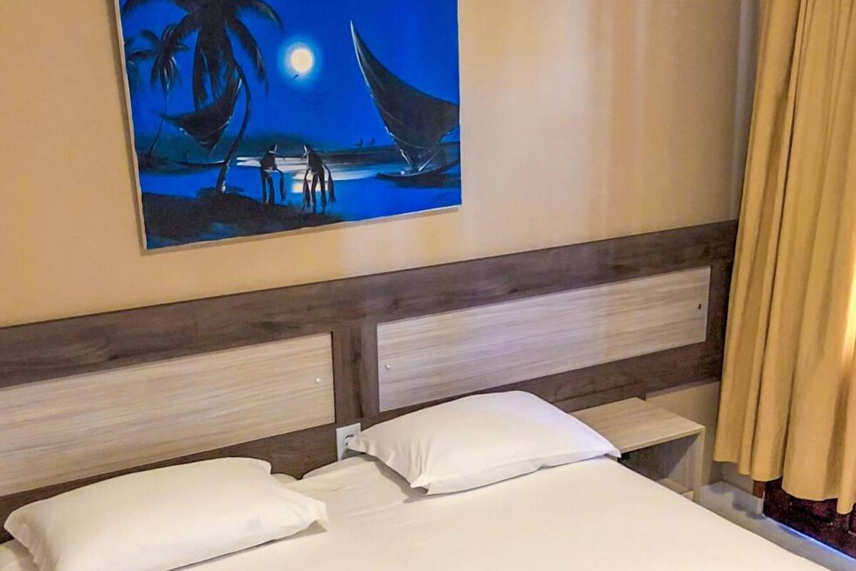 A hotel room with a bed, a headboard, a painting of a tropical scene, and a curtain.