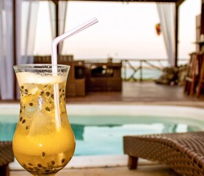 A glass of a yellow beverage with a straw, placed in front of a pool area with lounge chairs and a view of the pool. - Pousada Portal da Barra (Itarema)