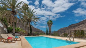 A swimming pool with lounge chairs, surrounded by palm trees and a mountainous backdrop. - GC0285 Holiday house with shared pool in Mogán (Mogán)