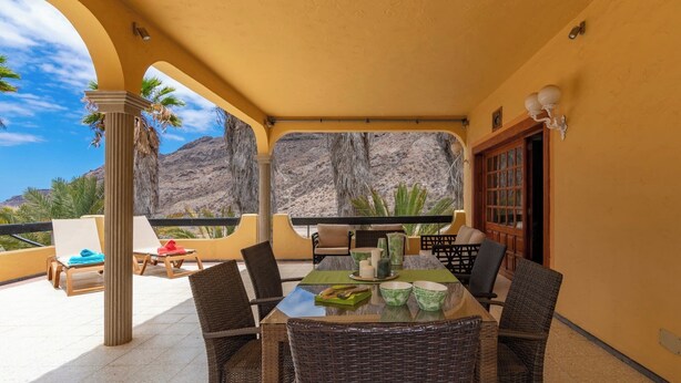 A covered outdoor seating area with wicker chairs and a glass table, overlooking a mountainous landscape. - GC0285 Holiday house with shared pool in Mogán (Mogán)