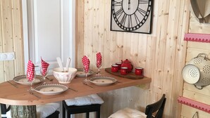 Een houten interieur met een ronde tafel gedekt voor vier personen, met wit en rood servies, een grote wandklok en een rustieke inrichting, waaronder een boomstam en houten wandbekleding.