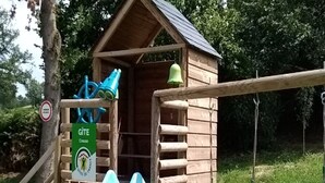 Een houten speelhuisje met een glijbaan, een bel en een bordje dat een Gîte camping aangeeft.