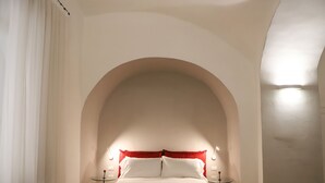 Une chambre à coucher dotée d’un grand plafond en arc de cercle, d’un lit recouvert de draps blancs, de coussins rouges et de deux tables de chevet avec des lampes.