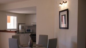 A dining area with a glass table and four chairs, a kitchen in the background, and a framed picture on the wall. - Lovely 2 bedrooms apartment in Torre Soli Nou (San Jaime Mediterráneo)