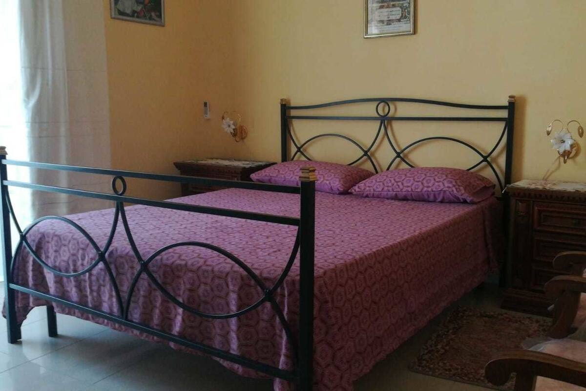 A single bed with a metal frame and a purple bedspread, a wooden nightstand, a small table with a statue, and a bench.