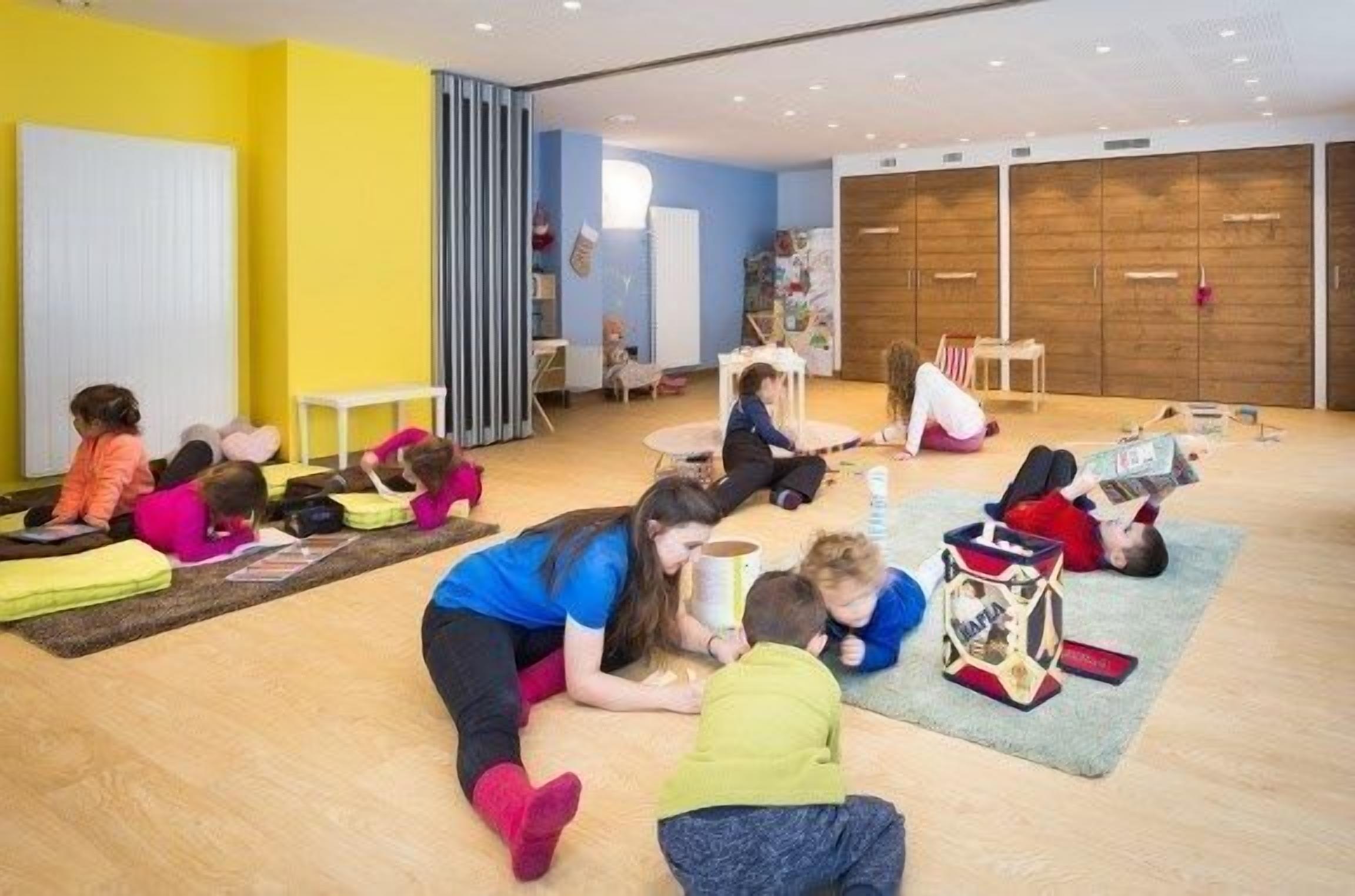 A brightly colored room with children and adults engaged in various activities, including reading and playing.