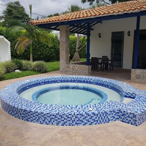 Ein kreisförmiger Pool mit blauen Mosaikfliesen, umgeben von einer Terrasse mit Fliesenboden und einer überdachten Sitzgelegenheit mit Tischen und Stühlen.