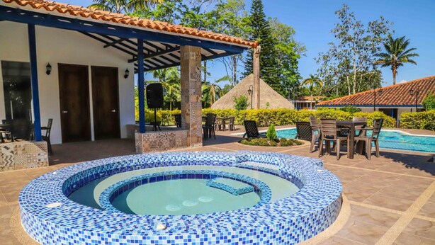 A circular pool with blue mosaic tiles, a covered patio area with chairs, and a thatched-roof structure in the background. - Hotel Campestre Santa Lucia (Montenegro)