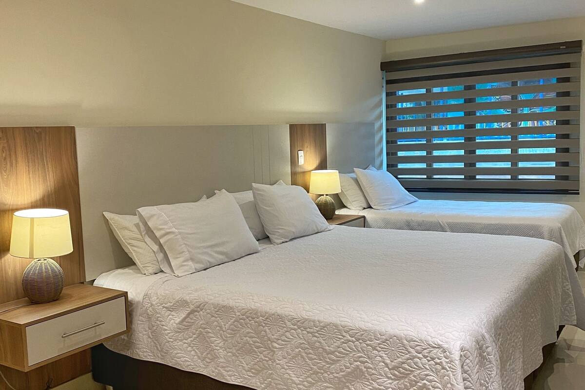 Ein Hotelzimmer mit zwei Betten, einem Nachttisch mit Lampe, einem Fenster mit Jalousien und einer Deckenleuchte.