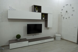 A modern living room with a flat-screen TV, built-in shelving, a small potted plant, and a wall clock. - A stone's throw from downtown and the sea modern comfort and the scent of Marsala bread (Marsala)