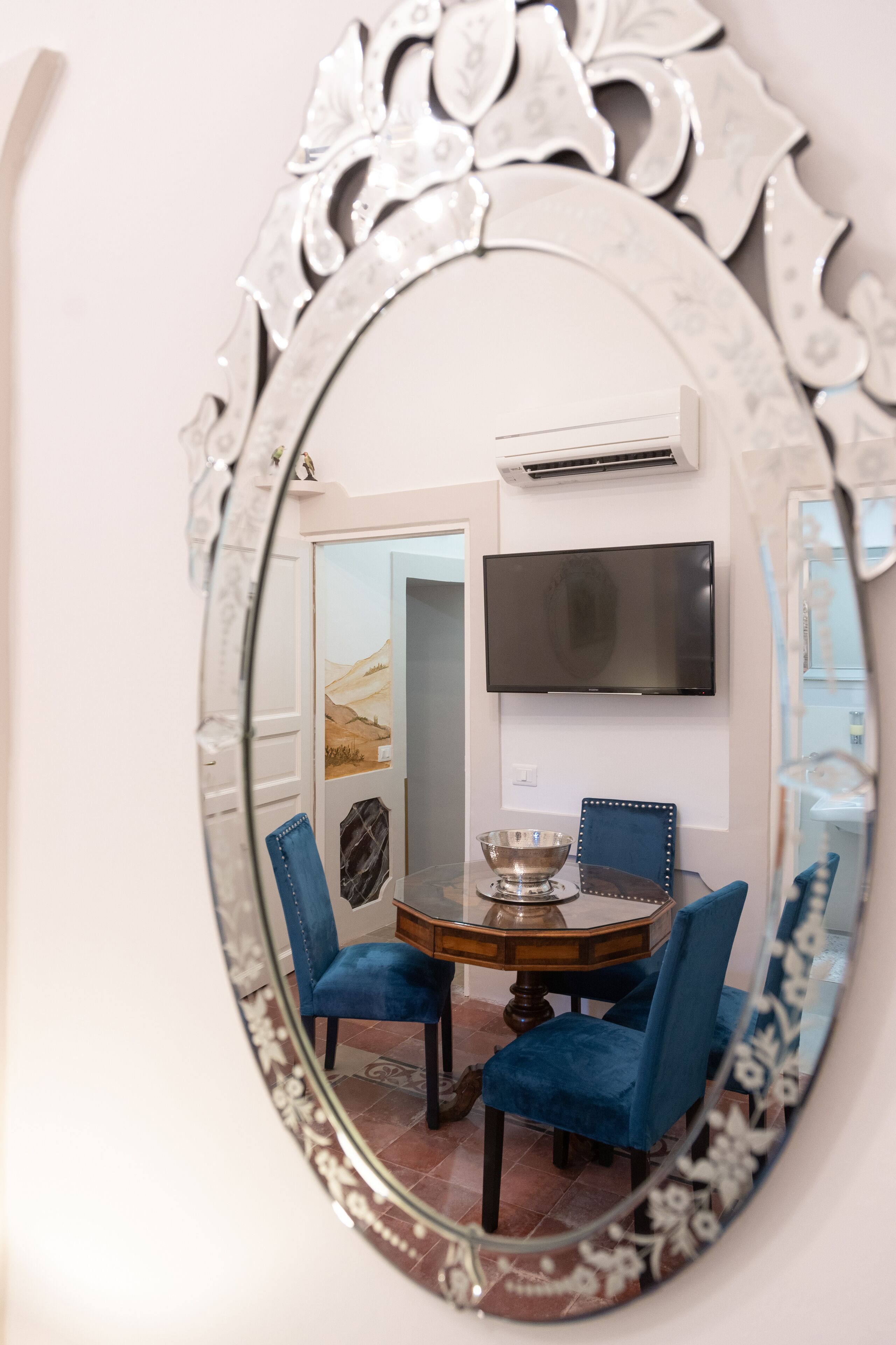 Un miroir ovale orné de motifs floraux, qui reflète un coin repas avec une table ronde, des chaises bleues et un téléviseur à écran plat fixé au mur.