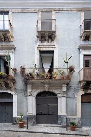 Une façade d’immeuble avec une entrée centrale, des balcons ornés de plantes en pot et une porte grise.
