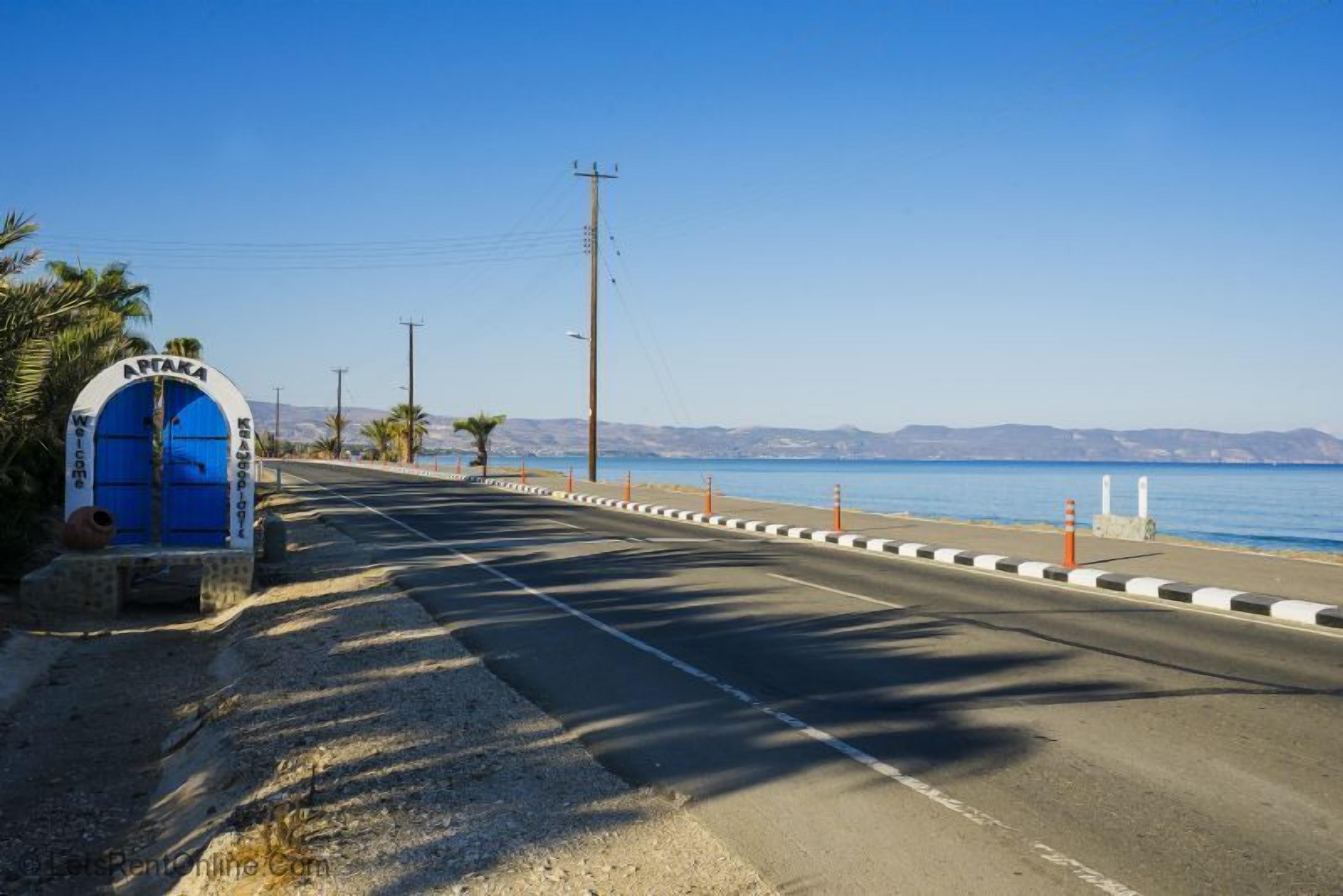 A coastal road with a blue entrance gate, palm trees, and a clear view of the sea.