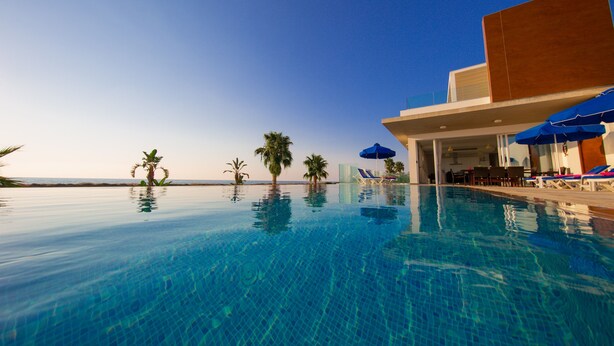 Ein klarblauer Infinity-Pool mit Blick auf den Ozean, Palmen und ein modernes Gebäude mit Liegestühlen und Sonnenschirmen.