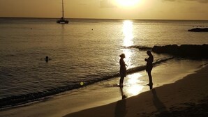 A beach scene with two people near the water's edge, a sailboat in the distance, and the sun setting over the horizon. - Seabreeze Villa: Best rates. Charming, bright & spacious. Truly a home from home (Atlantic Shores)
