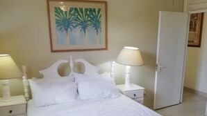 A bedroom with a bed, two bedside tables, a nightstand, a door, and a wall-mounted painting. - Seabreeze Villa: Best rates. Charming, bright & spacious. Truly a home from home (Atlantic Shores)