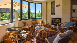A cozy living room with wicker furniture, a fireplace, and large windows offering a view of greenery. - Loire Valley with pool (Thizay)