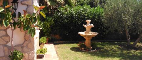 Ein Garten mit Steinmauer, Pflasterterrasse, Springbrunnen und verschiedenen Kübelpflanzen.