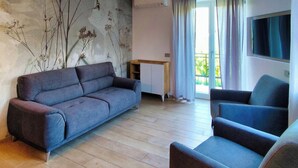 A modern living room with a grey sofa, a smaller armchair, a flat-screen TV mounted on the wall, and a window with curtains.