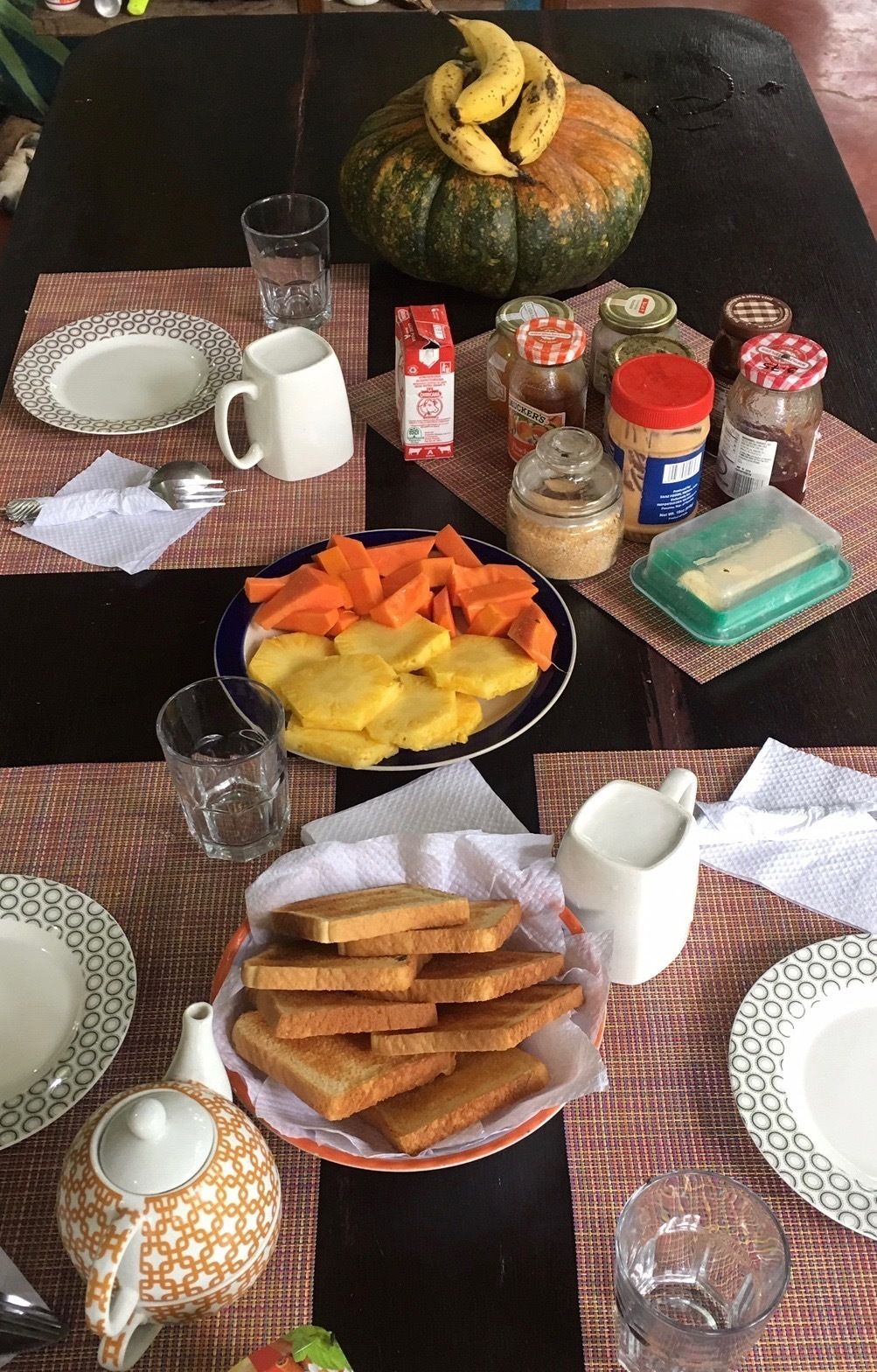 Una mesa dispuesta con platos, una jarra, un vaso, un bol de fruta, una cesta de tostadas, salsas y un calabacín.