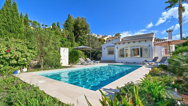 Piscina com espreguiçadeiras, rodeada por vegetação exuberante e um edifício branco com telhado em azulejo.