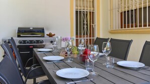 Uma área de refeições ao ar livre com uma mesa de madeira posta para quatro pessoas, rodeada por cadeiras. Uma churrasqueira é visível ao fundo, e uma janela com grades está à direita.