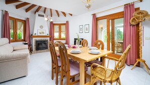 Uma sala de jantar com uma mesa de madeira posta para quatro pessoas, rodeada por cadeiras de madeira. Há um sofá e uma lareira ao fundo.
