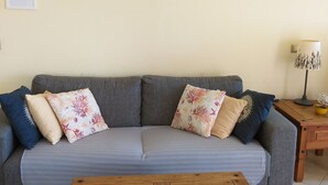 Ein Wohnzimmer mit grauem Sofa, floralen Kissen, einem Couchtisch aus Holz mit Meeresschnecken, einem an der Wand befestigten Globus und einem gerahmten Bild.