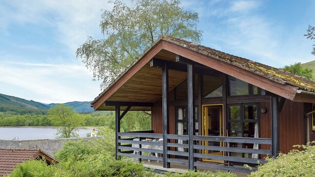 A wooden cabin with a balcony overlooking a lake, surrounded by greenery.