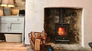 A cozy room with a stone wall, a fireplace with a black stove, a lamp, a basket of firewood, and a framed map on the wall.