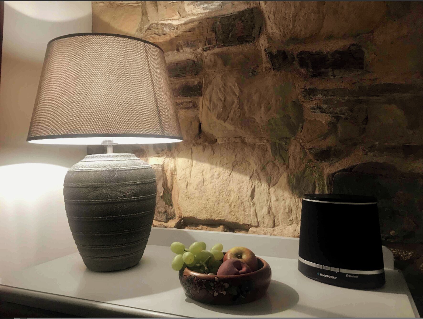 Une lampe, un saladier rempli de fruits et un récipient noir sur une table, le tout adossé à un mur de pierre.