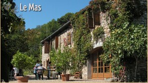 Edificio de piedra con puerta y ventanas de madera, rodeado de vegetación y gente.