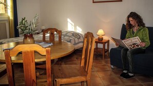 Una sala de estar acogedora con una mesa redonda de madera, dos sillas de madera, un sofá azul, una mesita auxiliar con una lámpara y una planta en maceta.