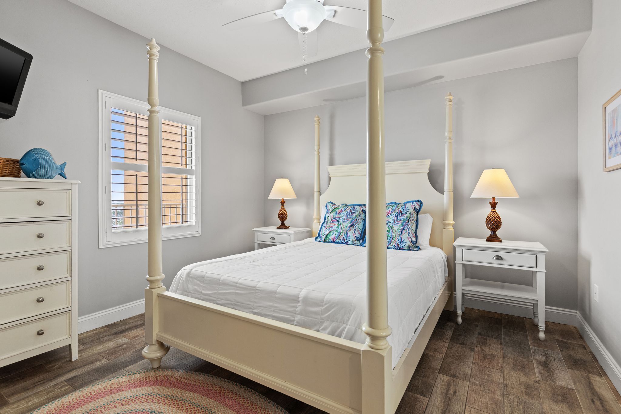 A four-poster bed with white bedding, two bedside tables with lamps, a dresser, and a television on the left wall.