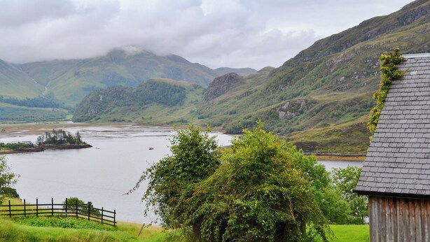 A rural landscape with a river, mountains, and a wooden building.