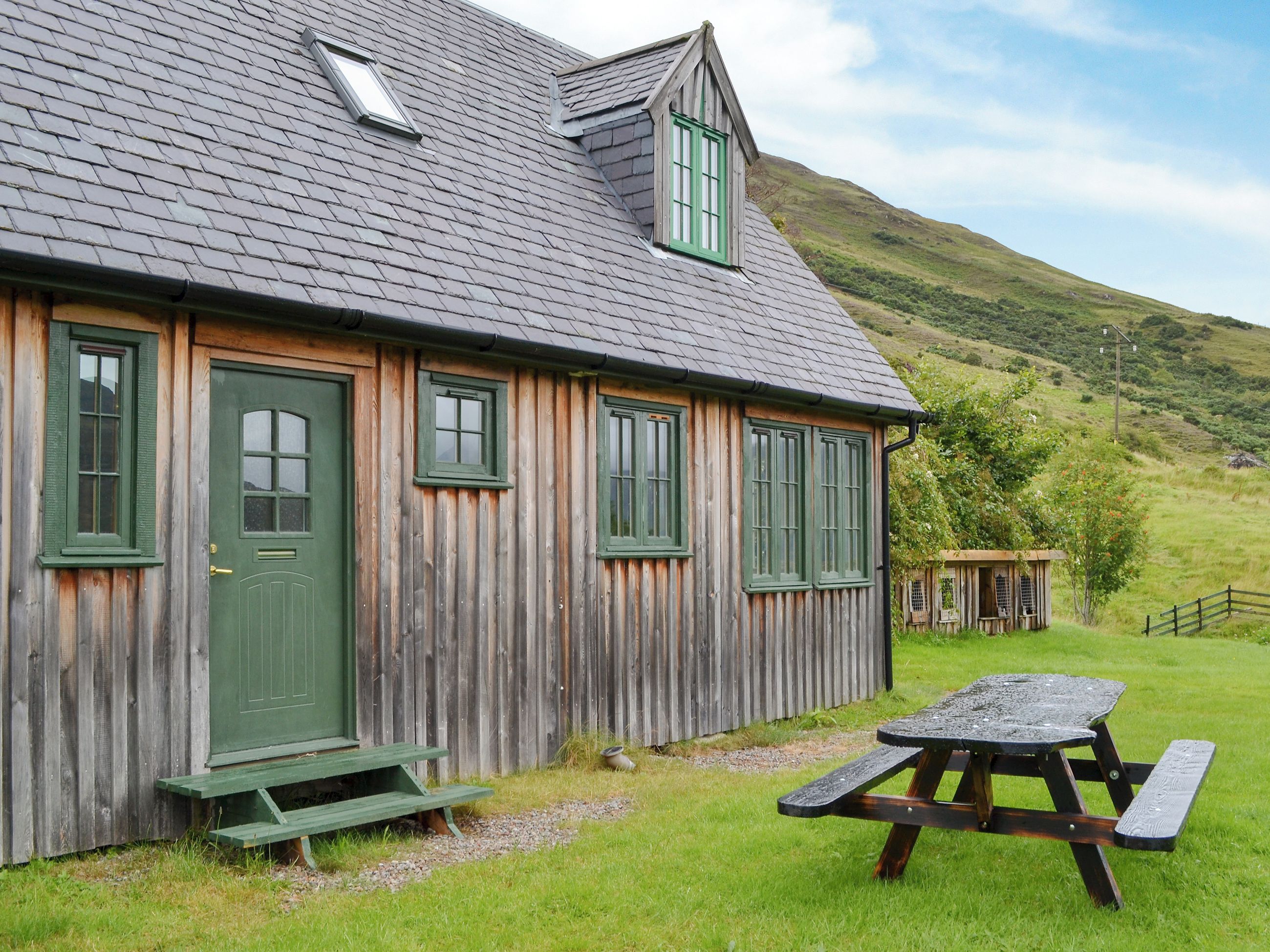 A wooden cottage with a green door and windows, a picnic table, and a small shed in a grassy area with a hill in the background.
