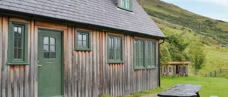 Una cabaña de madera con puerta y ventanas verdes, una mesa de picnic y un cobertizo pequeño en un área cubierta de césped, con una colina al fondo.