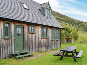 A wooden cottage with a green door and windows, a picnic table, and a small shed in a grassy area with a hill in the background. - 2 bedroom accommodation in Dornie (Dornie)