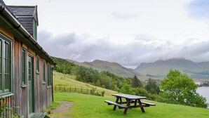 A rustic building with a wooden exterior and a picnic table in a grassy area with a view of mountains and a lake.