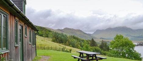 A rustic building with a wooden exterior and a picnic table in a grassy area with a view of mountains and a lake.
